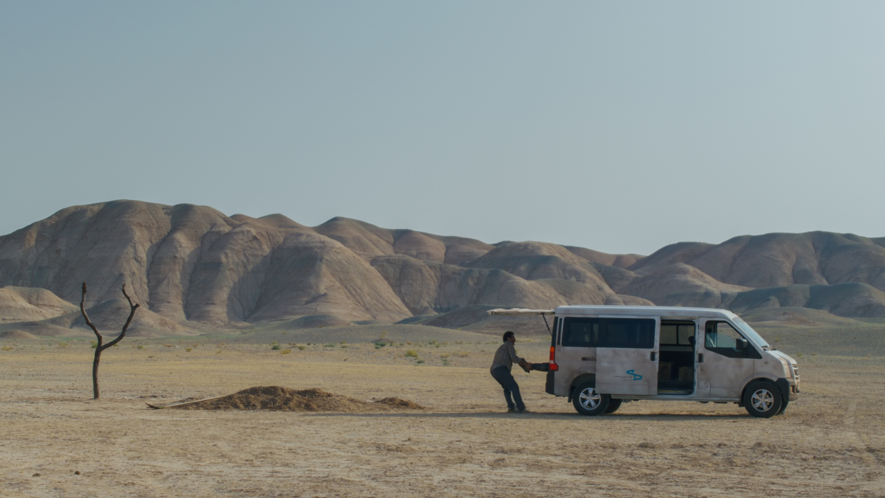 Um homem descarrega algo da traseira de um furgão no meio do deserto. Atrás dele tem um buraco, um monte de terra e uma pequena árvore.